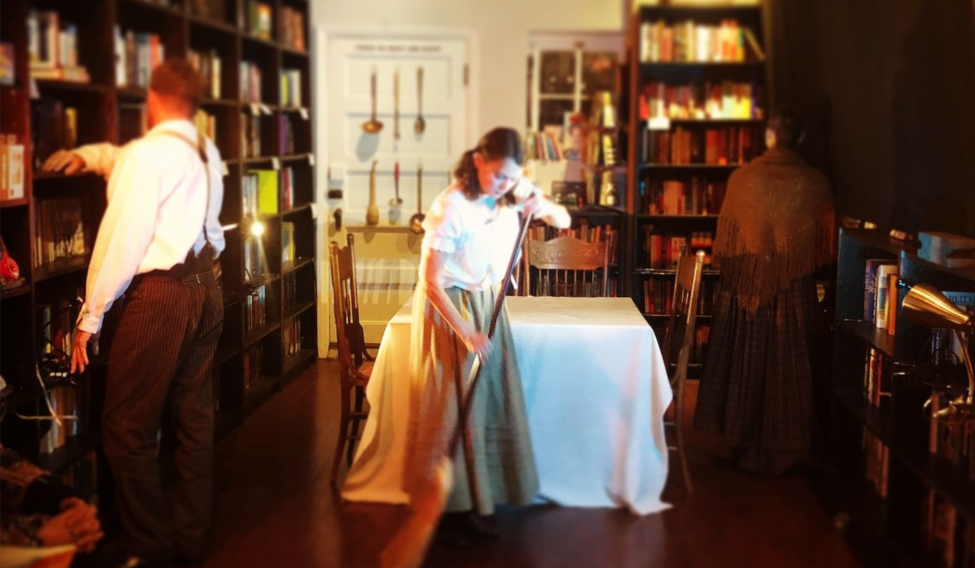 Julie sweeping the floor in front of a table, two other figures have their backs turned