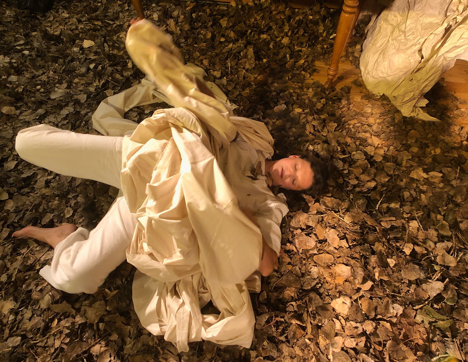 julie in white, covered in a sheet, laying face up in dead leaves