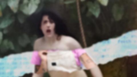 Truth emerging from her well, with the paper ripped across her bust, revealing white coveralls, a pink tshirt, and tatood arms. Text from a script is blurry in the background.