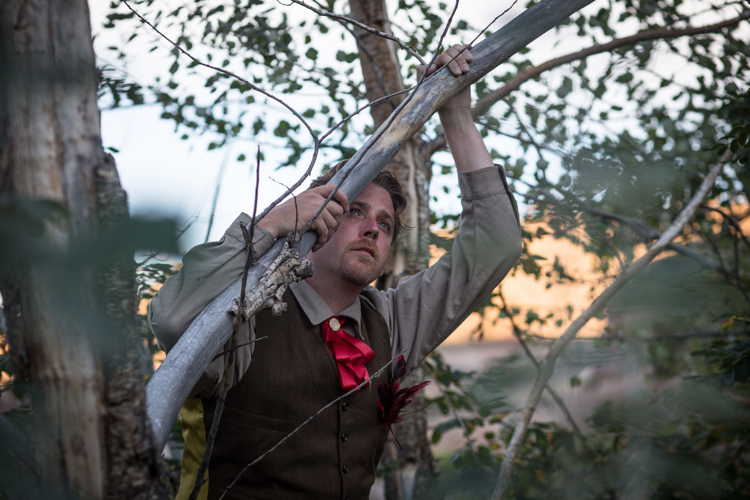 Kenny in a brown vest and red bow, leaning on a branch and looking into the distance