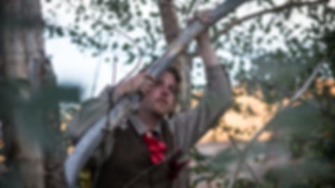 Kenny in a brown vest and red bow, leaning on a branch and looking into the distance