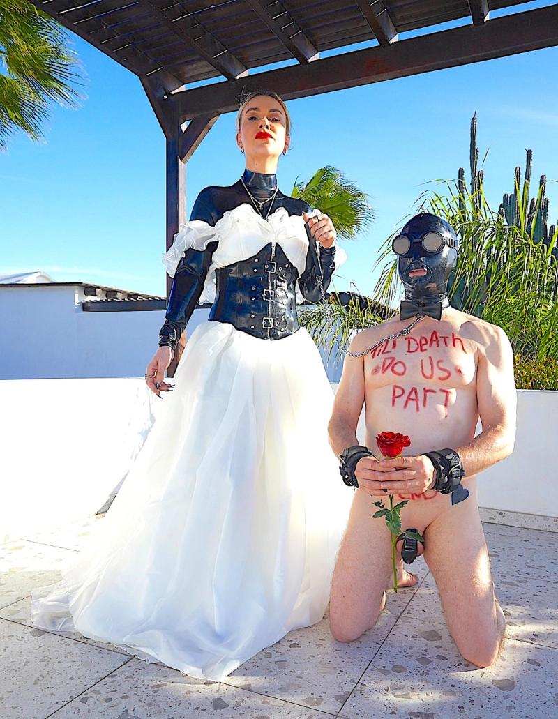 Свадебное фото Госпожи и раба. Госпожа одета в свадебное платье поверх латексного костюма. раб стоит на коленях в маске и поясе верности, он держит в руке розу