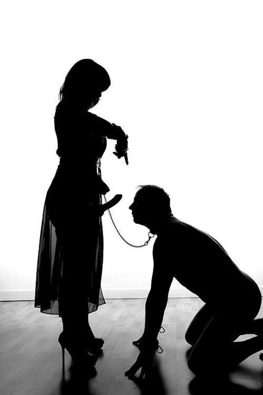 Девушка со страпоном указывает пальцем на него мужчине на коленях, как бы предлагая ему его пососать