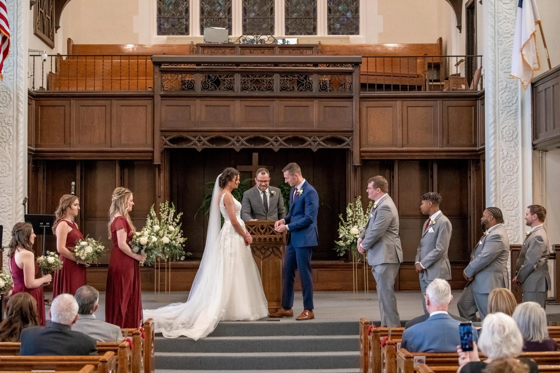 A bride and groom face each other at the chancel, their wedding party beside them and guests in the pews
