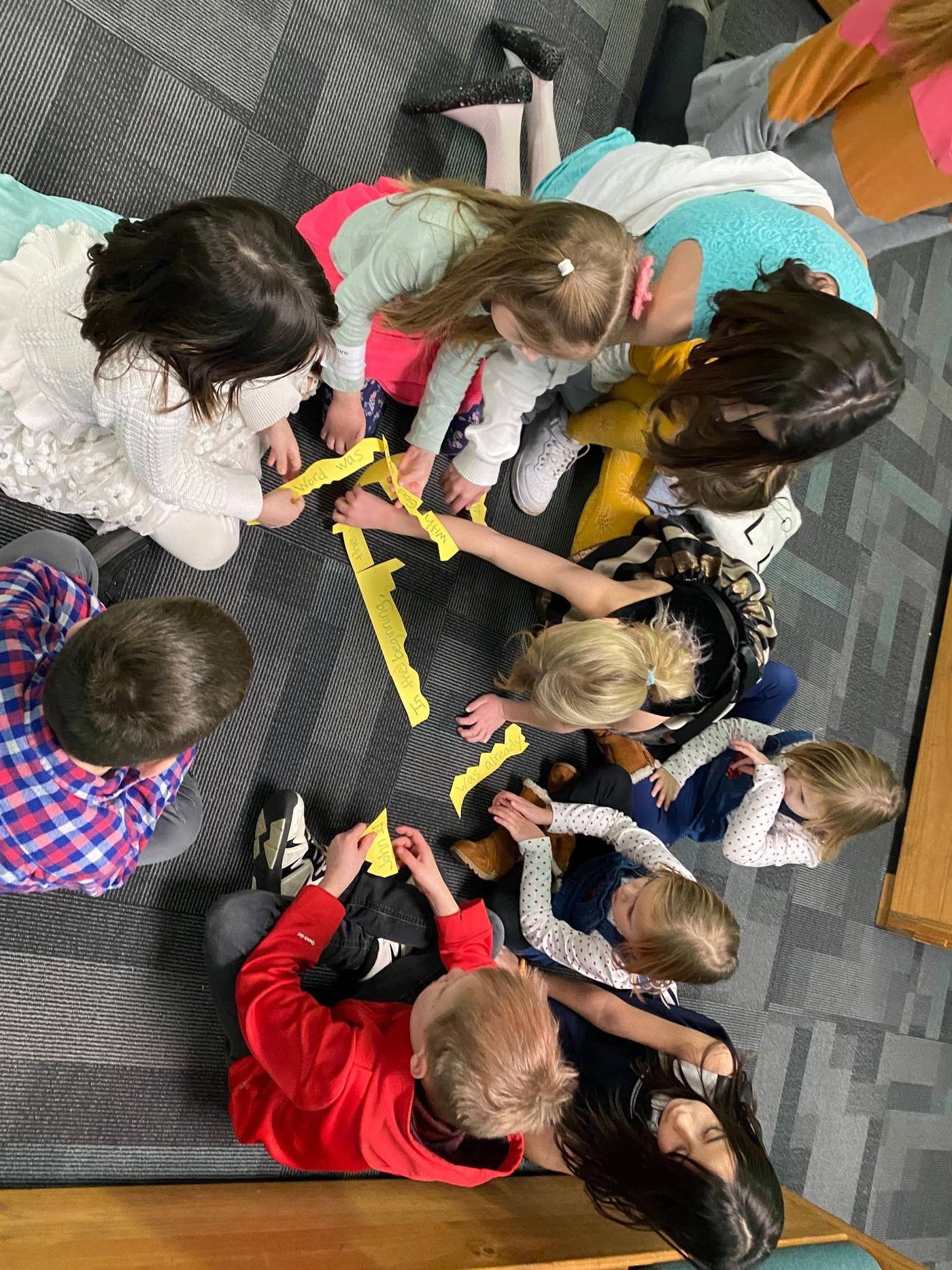 Children are seen from directly above, gathered around strips of yellow tape laid out on the floor for a group game.