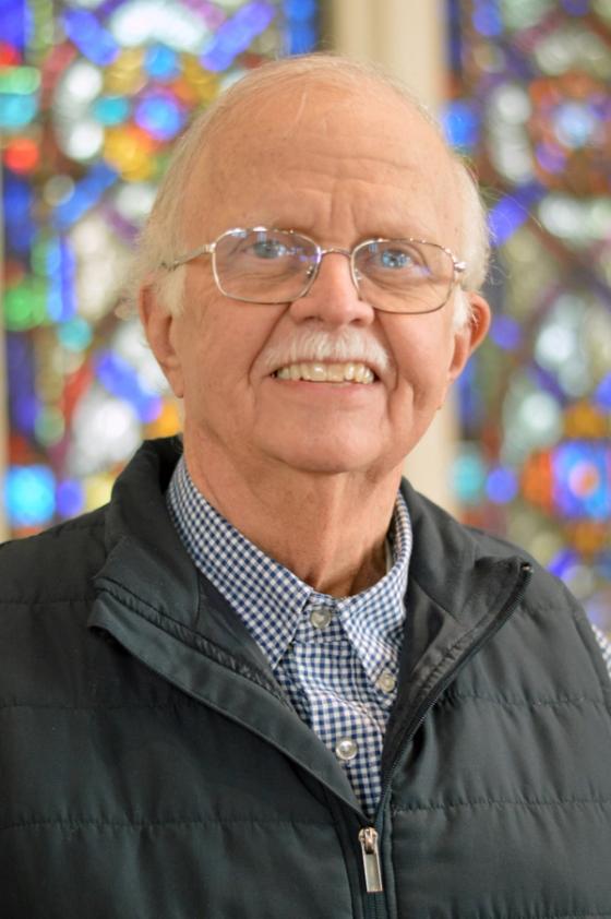Joe Songer, an older man with glasses, smiling in front of a stained glass window.