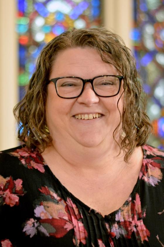 Sandi Brzak smiling, wearing glasses and a floral top, a stained glass window behind her.