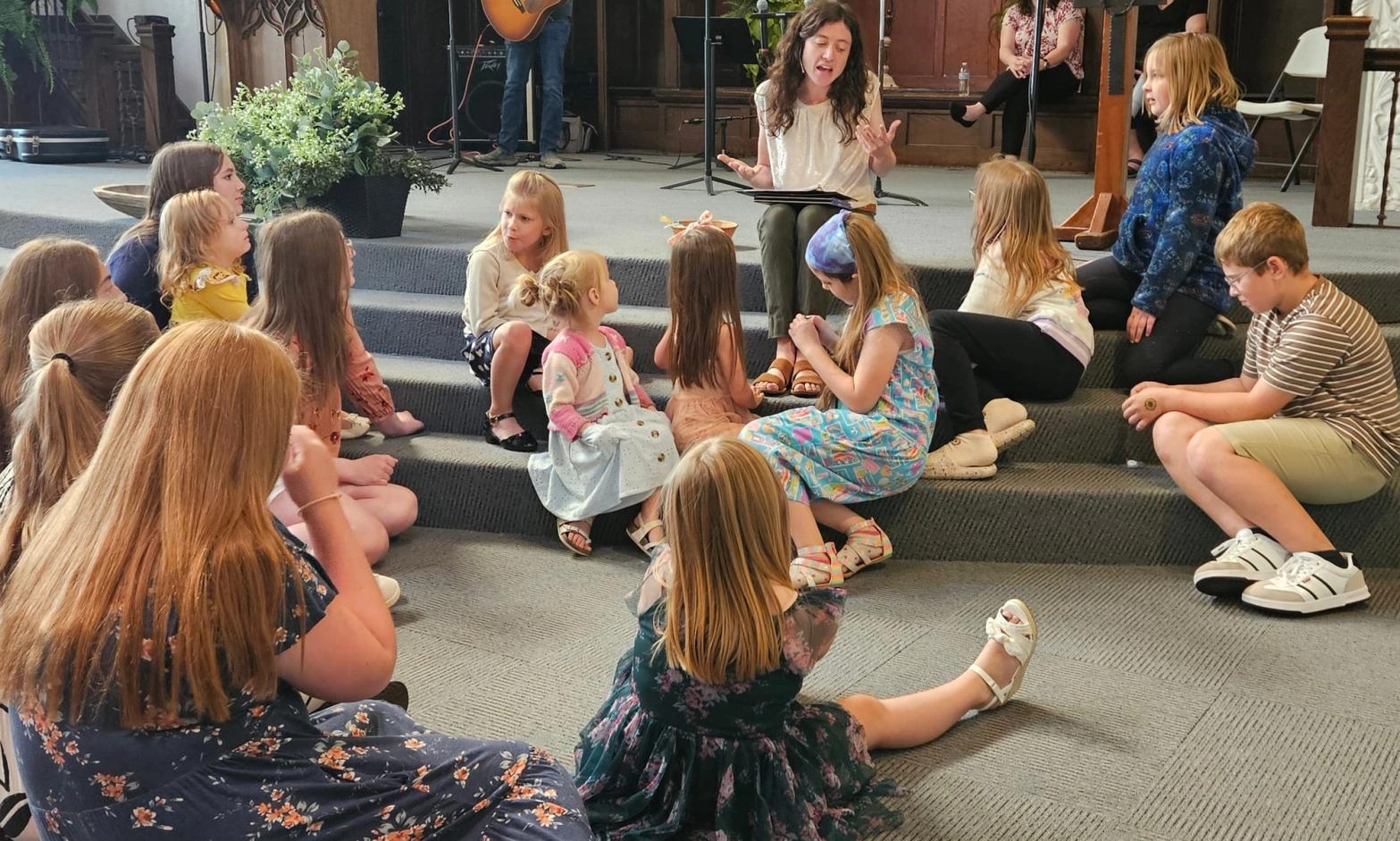 Children sit on the chancel steps around Kendall as she reads to them, at the blessing of the backpacks.