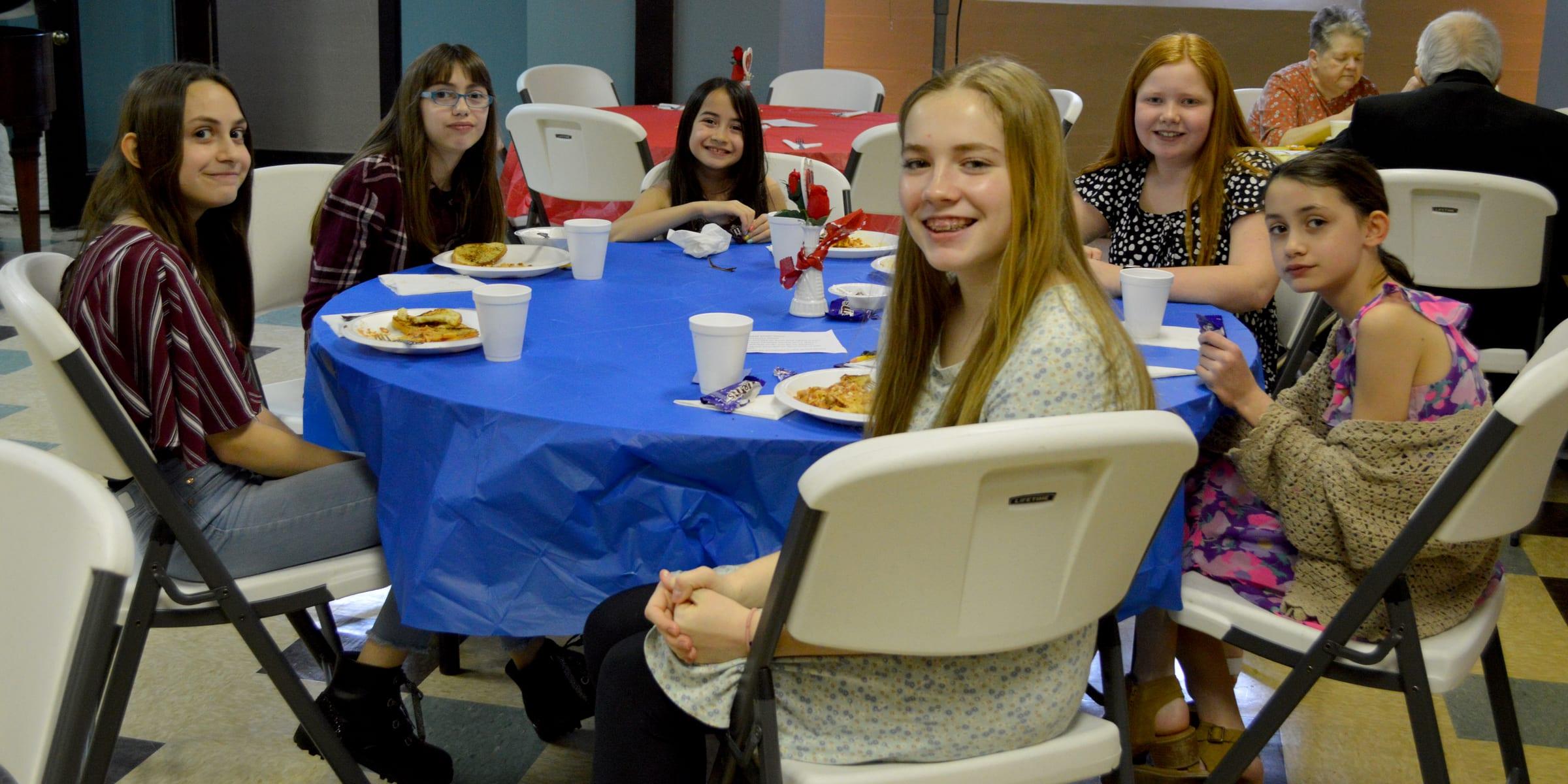A group of girls of different ages seated around a round table set with a blue tablecloth, plates and cups, smiling for the camera.