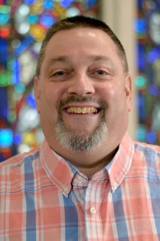 Ed Brzak smiling in a plaid shirt, a stained glass window behind him.