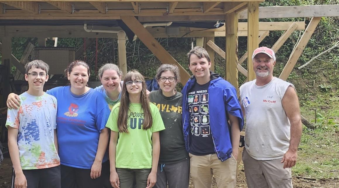 A mission trip group posed together under a wooden building frame they are constructing outdoors.
