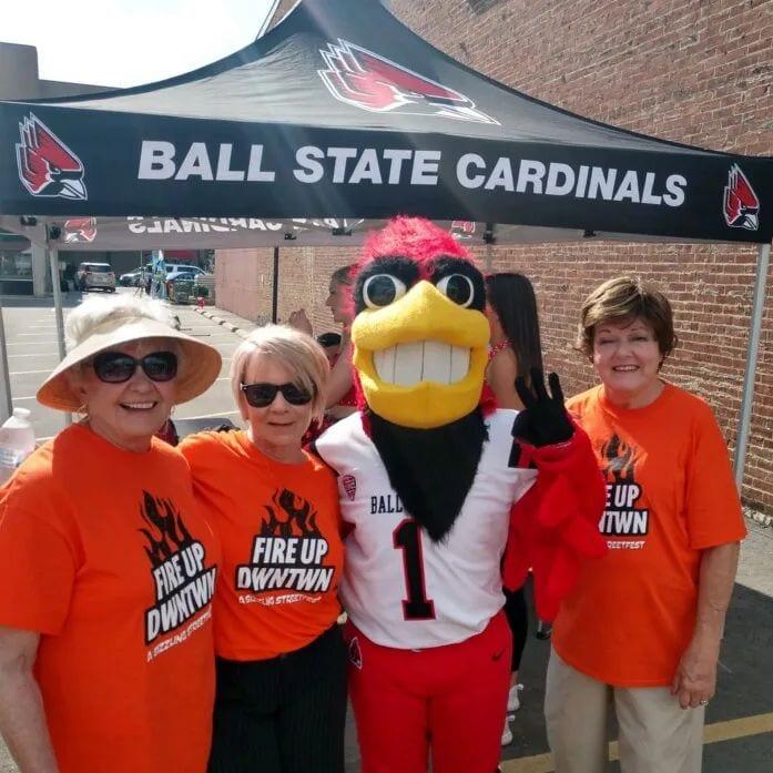 A group of women in orange shirts pose with the Ball State Cardinals mascot under a Ball State tent at an outdoor event.