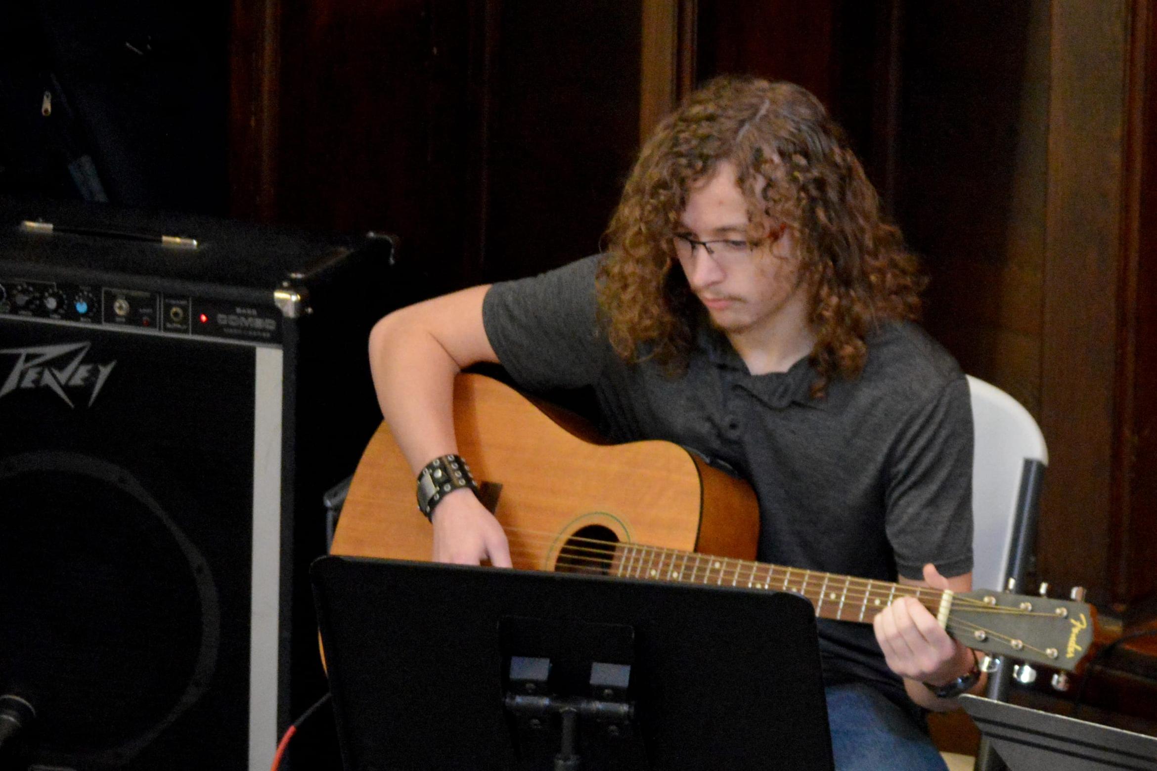 A young man with curly hair playing an acoustic guitar, reading from a music stand, an amplifier beside him.