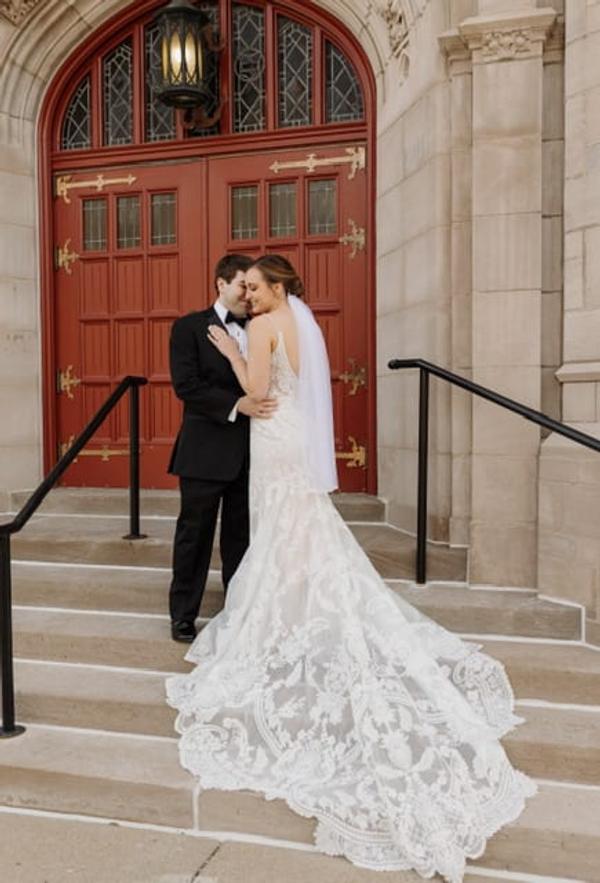 A bride and groom embrace on the front steps before the church's red doors, her lace train spread down the stairs