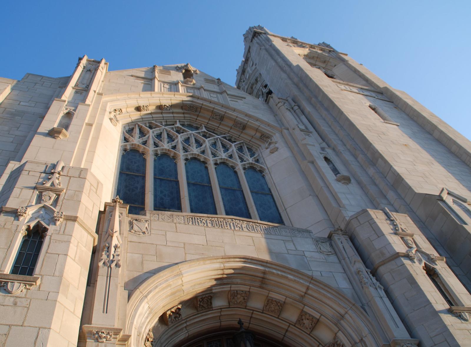 The limestone church facade rises steeply with a large arched stained glass window and the bell tower above, against a clear blue sky.