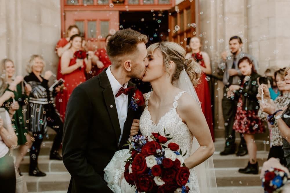 A bride and groom kiss on the church steps while their wedding party blows bubbles
