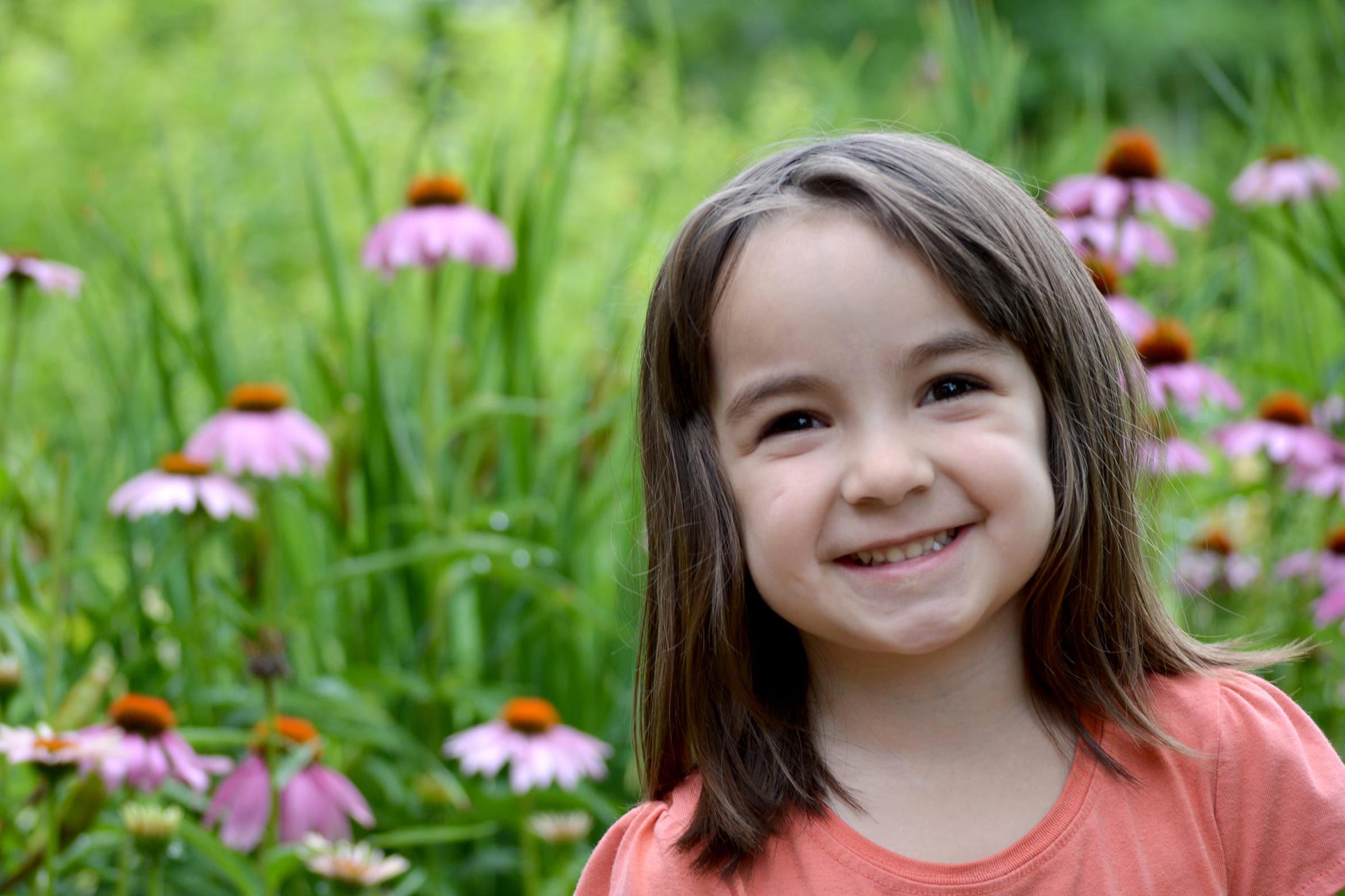 A young girl smiles outdoors, standing among tall purple coneflowers in a garden.