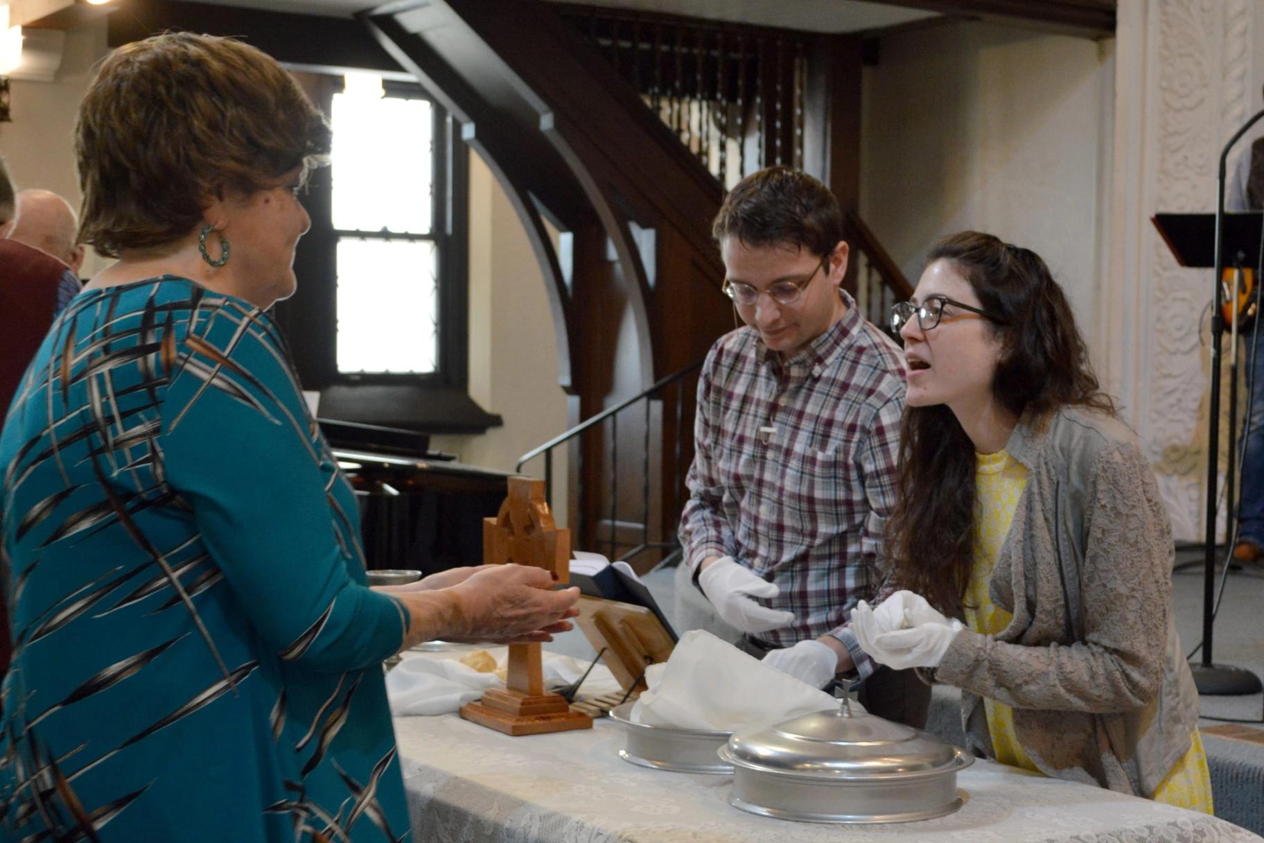 Two women and a man handle silver communion trays and a cloth at a table, preparing the elements.