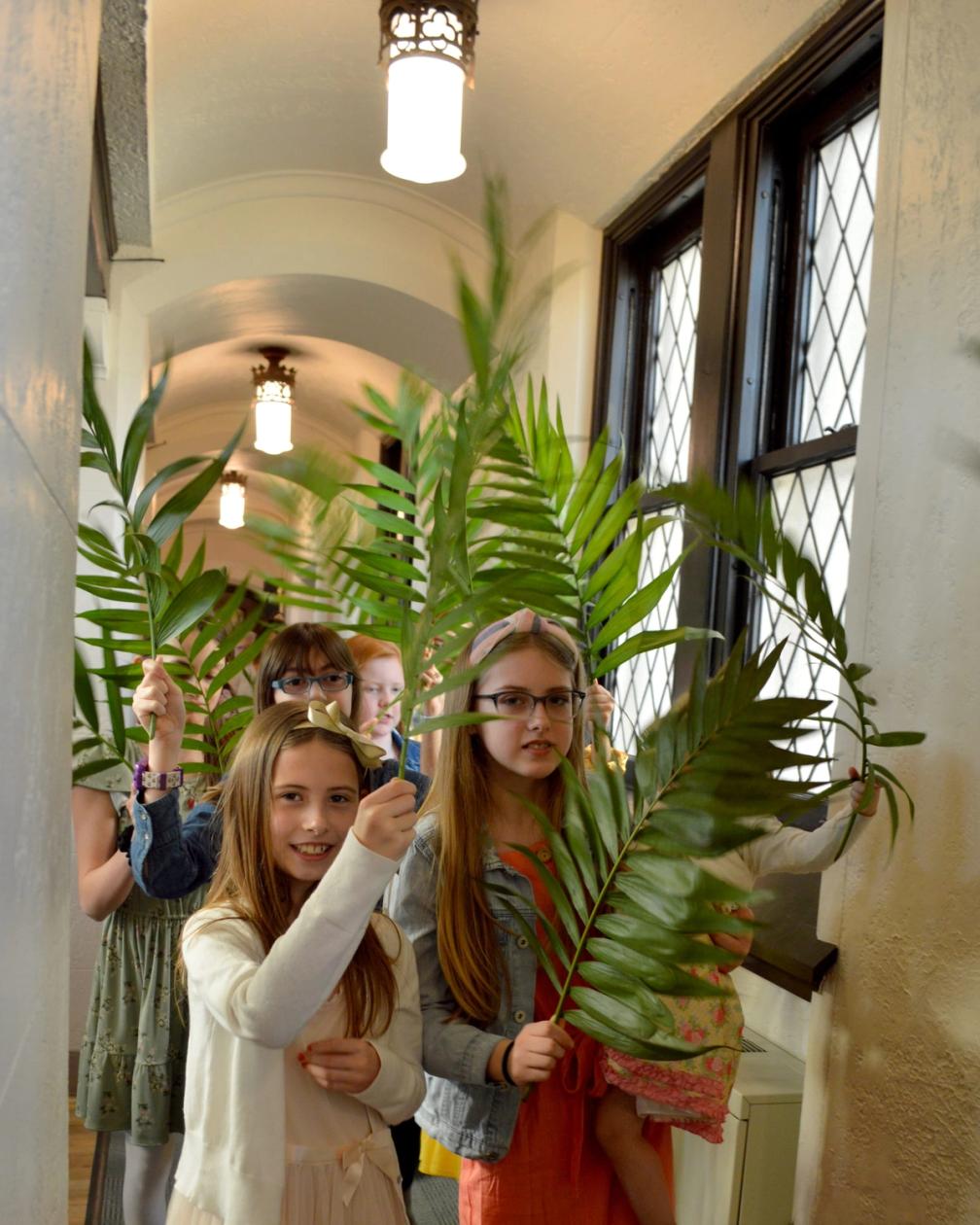 Children holding palm branches in a stone hallway, warm light coming through the leaded windows.
