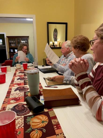 A group of adults sit together at a table set with an autumn runner and papers, talking and laughing during a fall dinner.