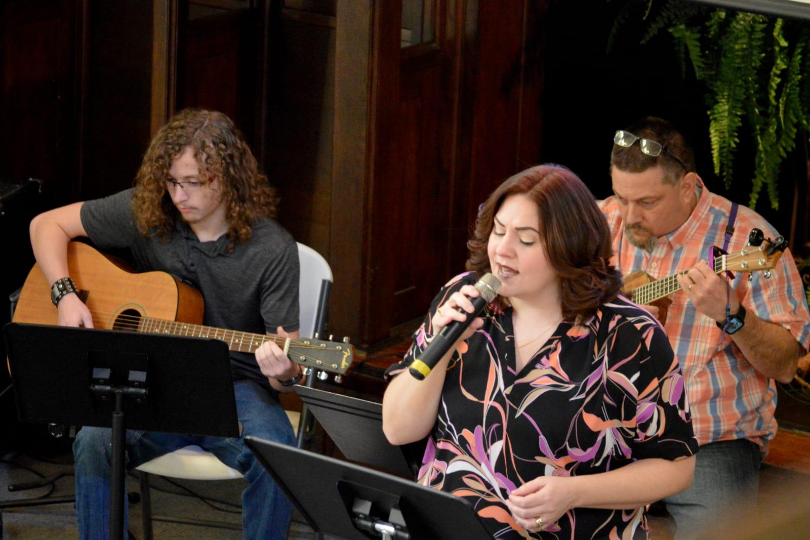 A guitarist, a singer holding a microphone, and a second guitarist perform together indoors.