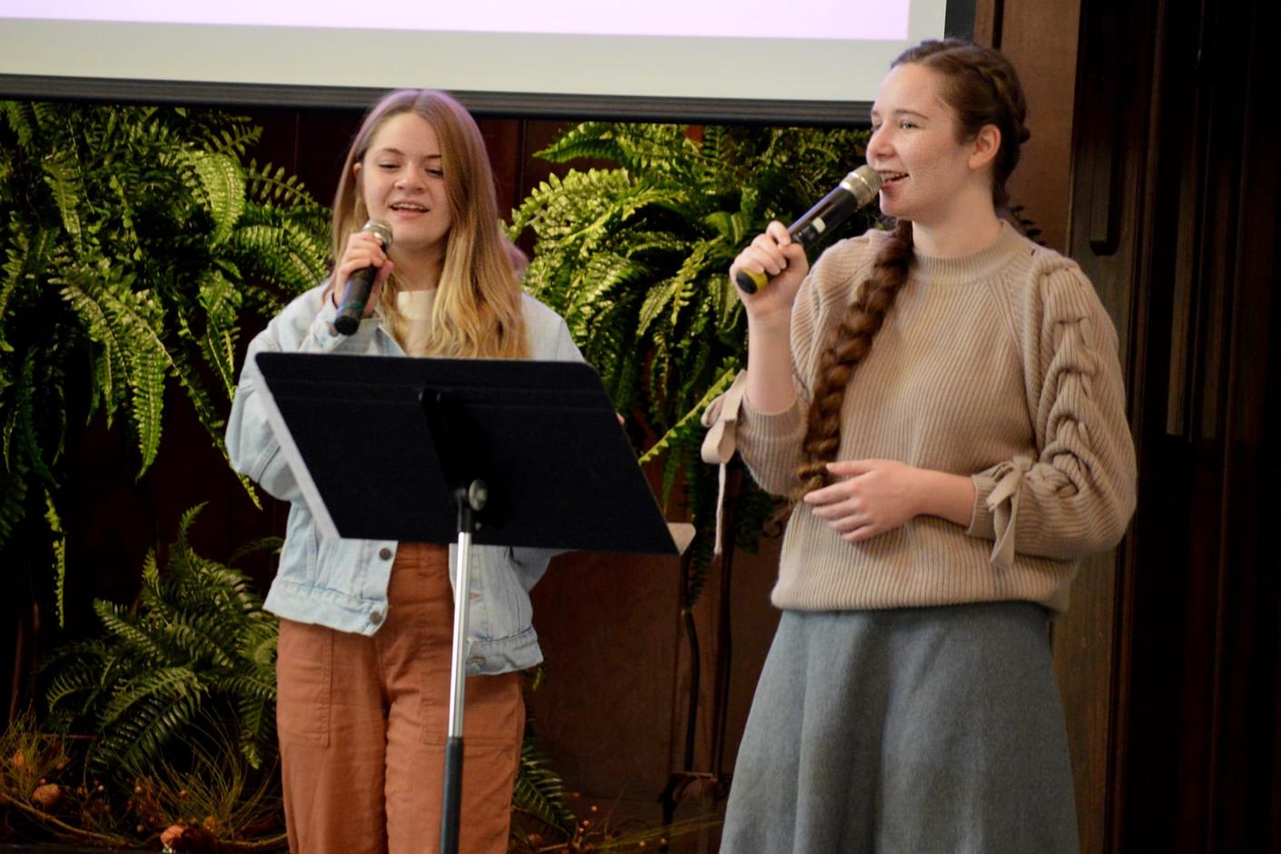 Two teenage girls sing together into microphones at an indoor event, with greenery behind them.
