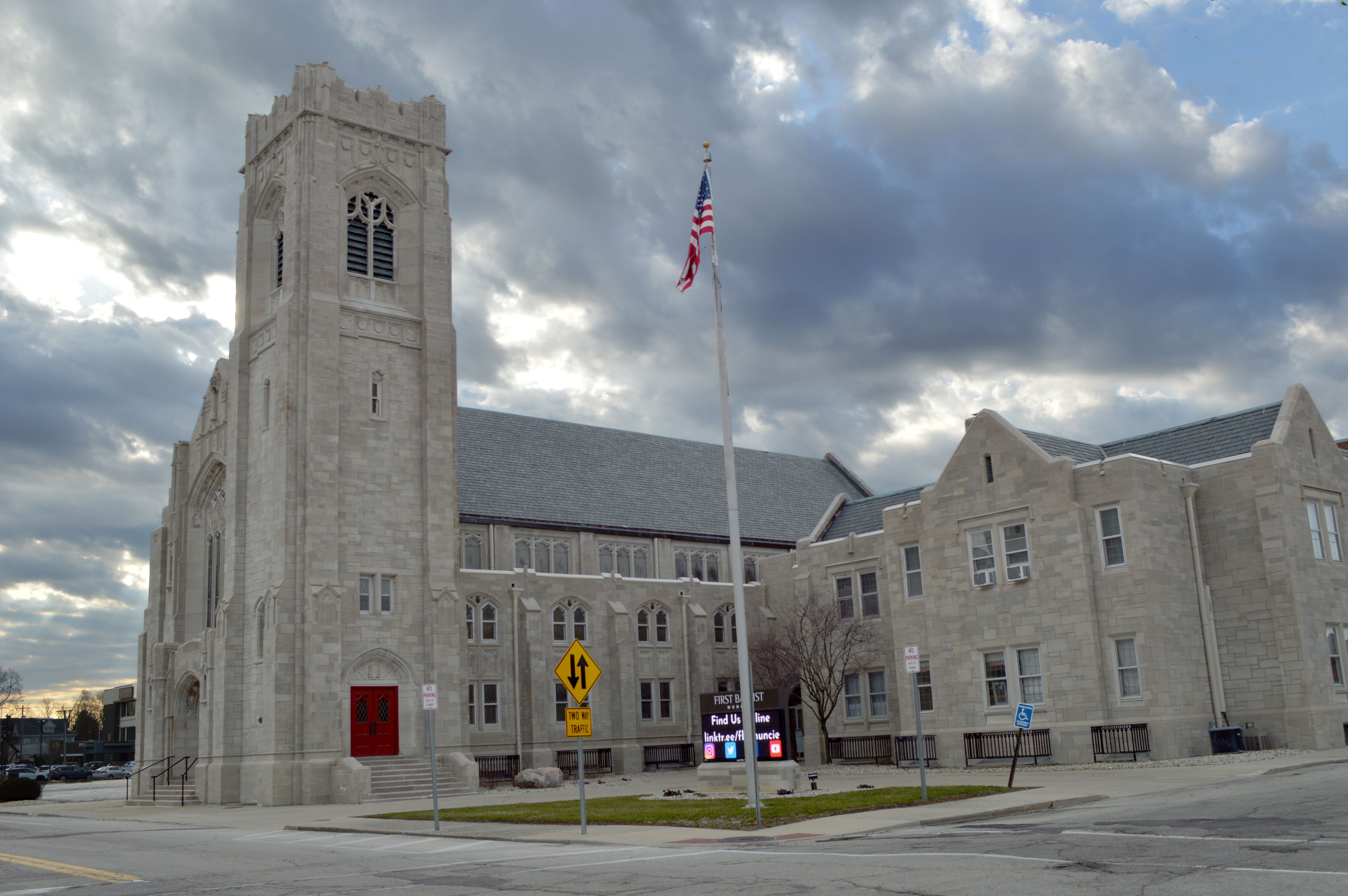 The whole church at dusk under a cloudy sky: the limestone bell tower with its red door, the flag, the lit sign on the lawn and the long wing beside it
