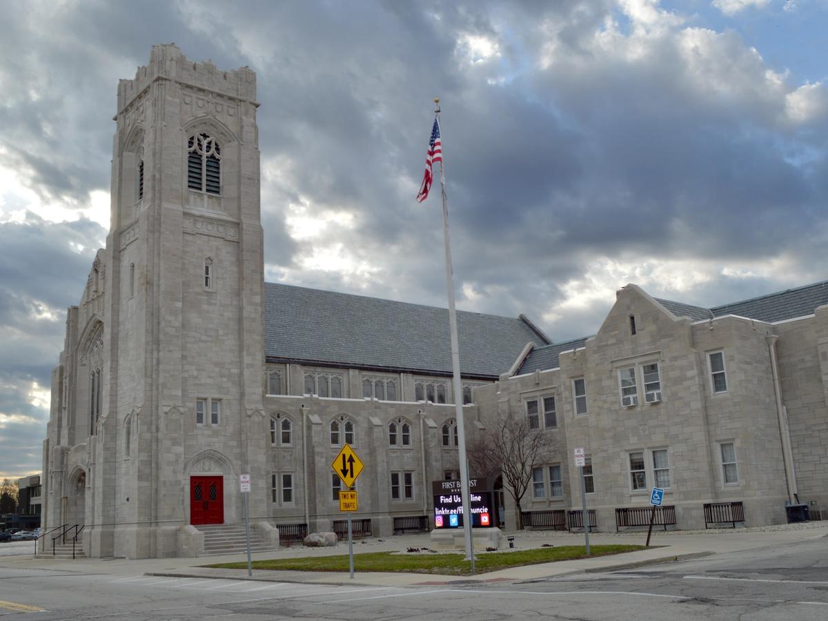 The whole church at dusk under a cloudy sky: the limestone bell tower with its red door, the flag, the lit sign on the lawn and the long wing beside it