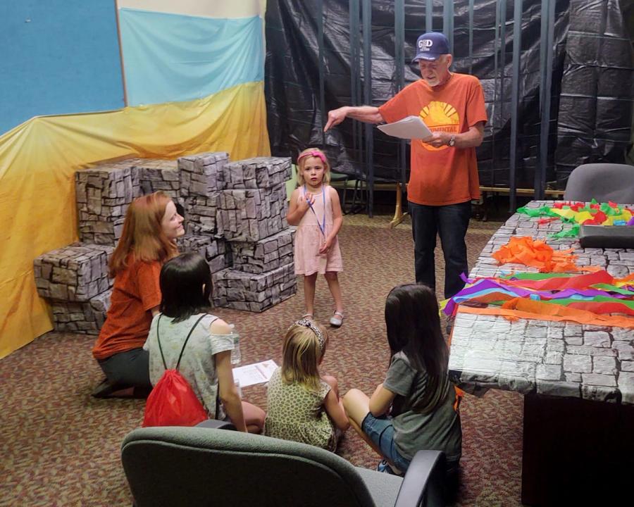 Children and adults gather around a display of blue and yellow decorated boxes and a fabric backdrop at an indoor event.