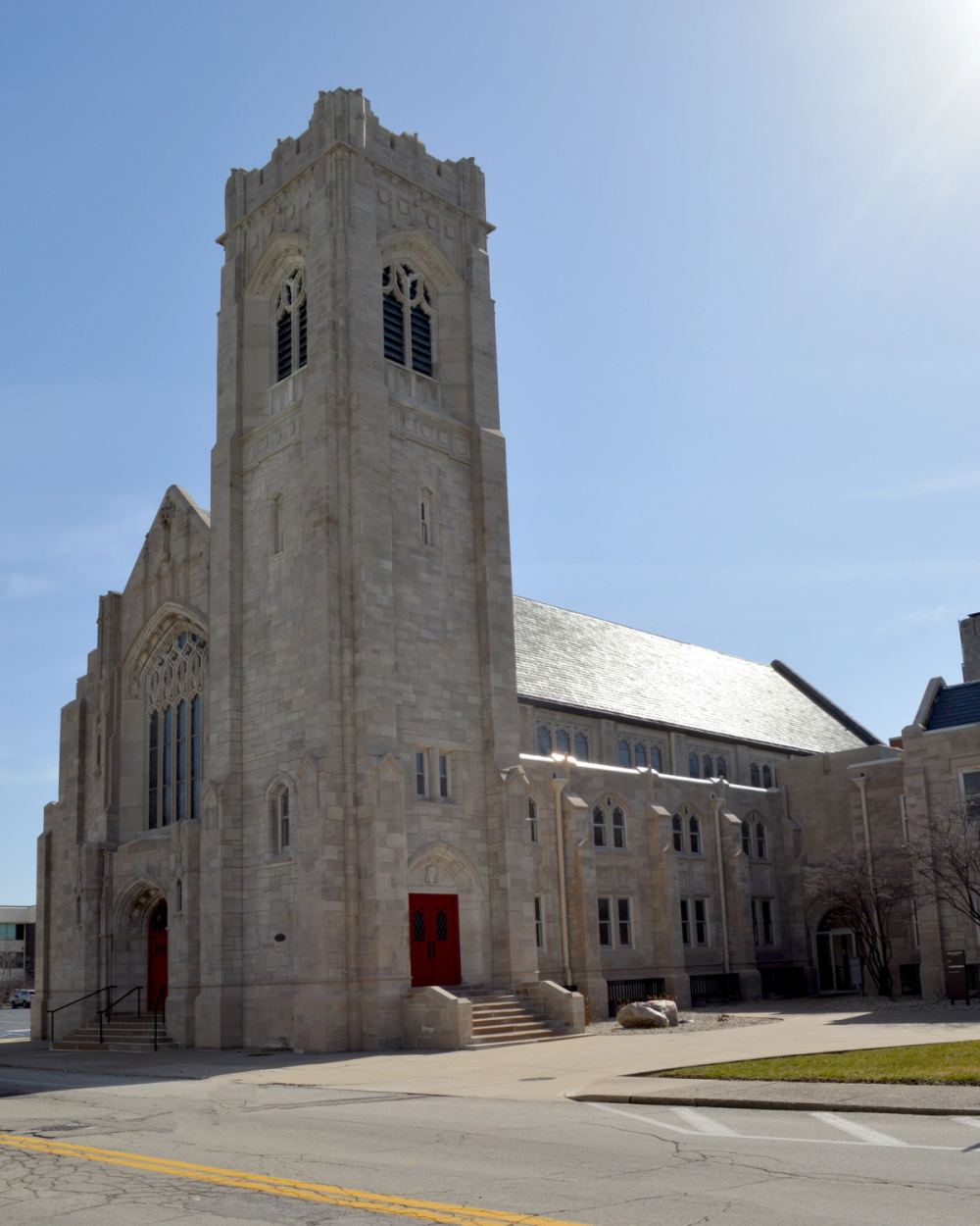 The full limestone church building is seen from a street corner on a sunny day, tower and wings visible.