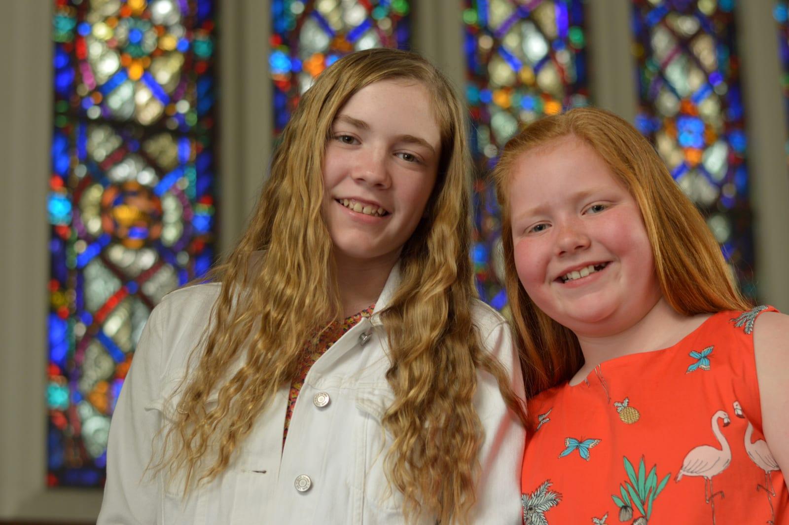 Two smiling girls, one with red hair, pose together in front of colorful stained glass windows.