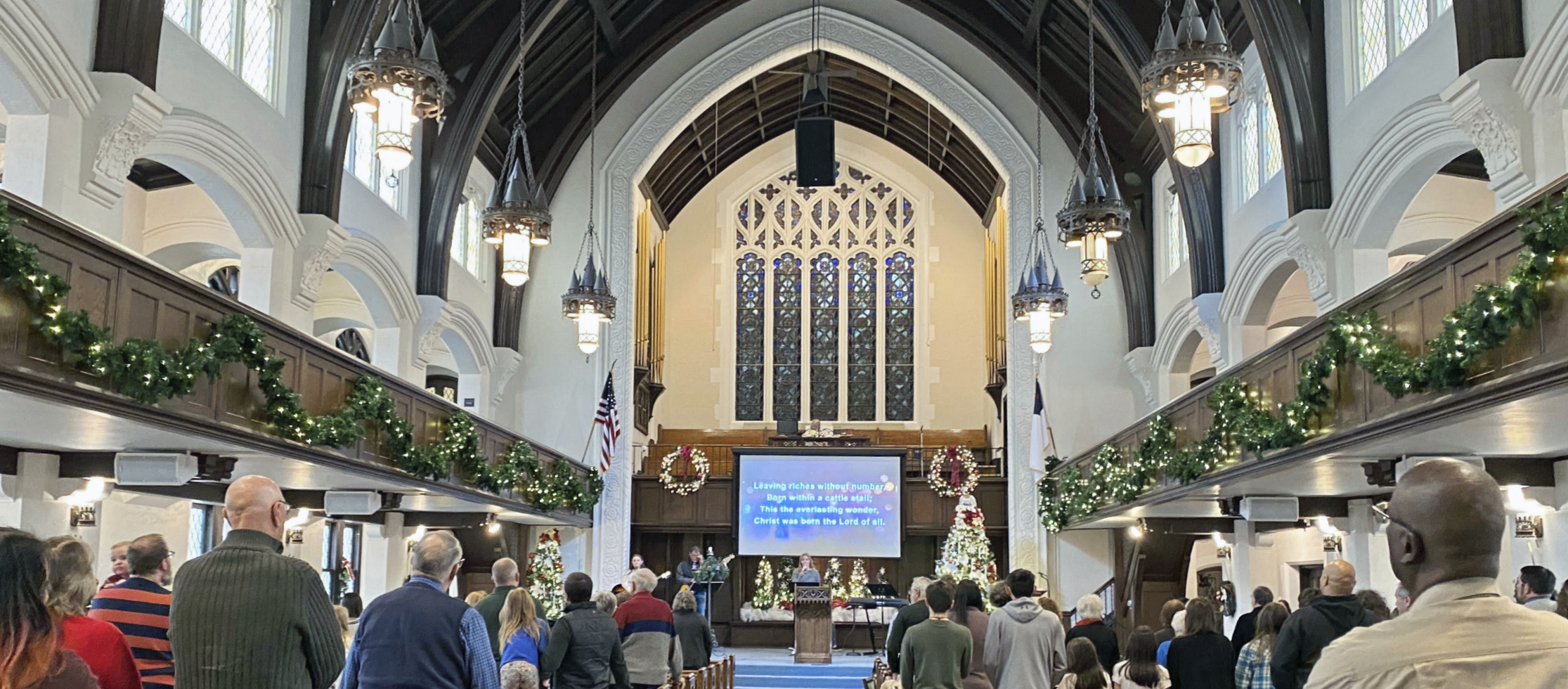 A high ceiling sanctuary decorated for Advent with Garland on the edge of the balcony, lights, and a stained glass window behind a pulpit adorned with lighted trees and wreaths, chandeliers hang from the ceiling.
