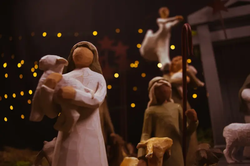 Wooden nativity scene with figures holding a lamb and shepherd's staff. Background features warm, glowing lights and dark, starry setting.