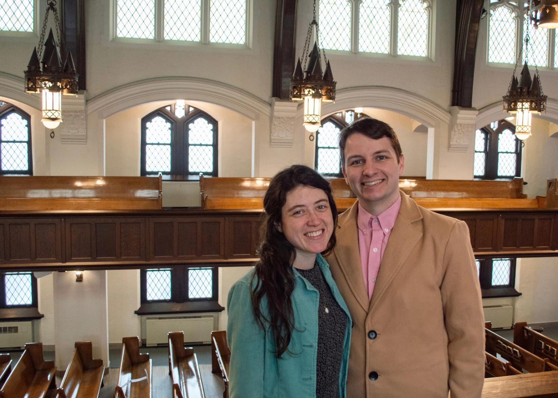 A man and a woman smiling together in the sanctuary balcony, arched windows and pews visible behind them.