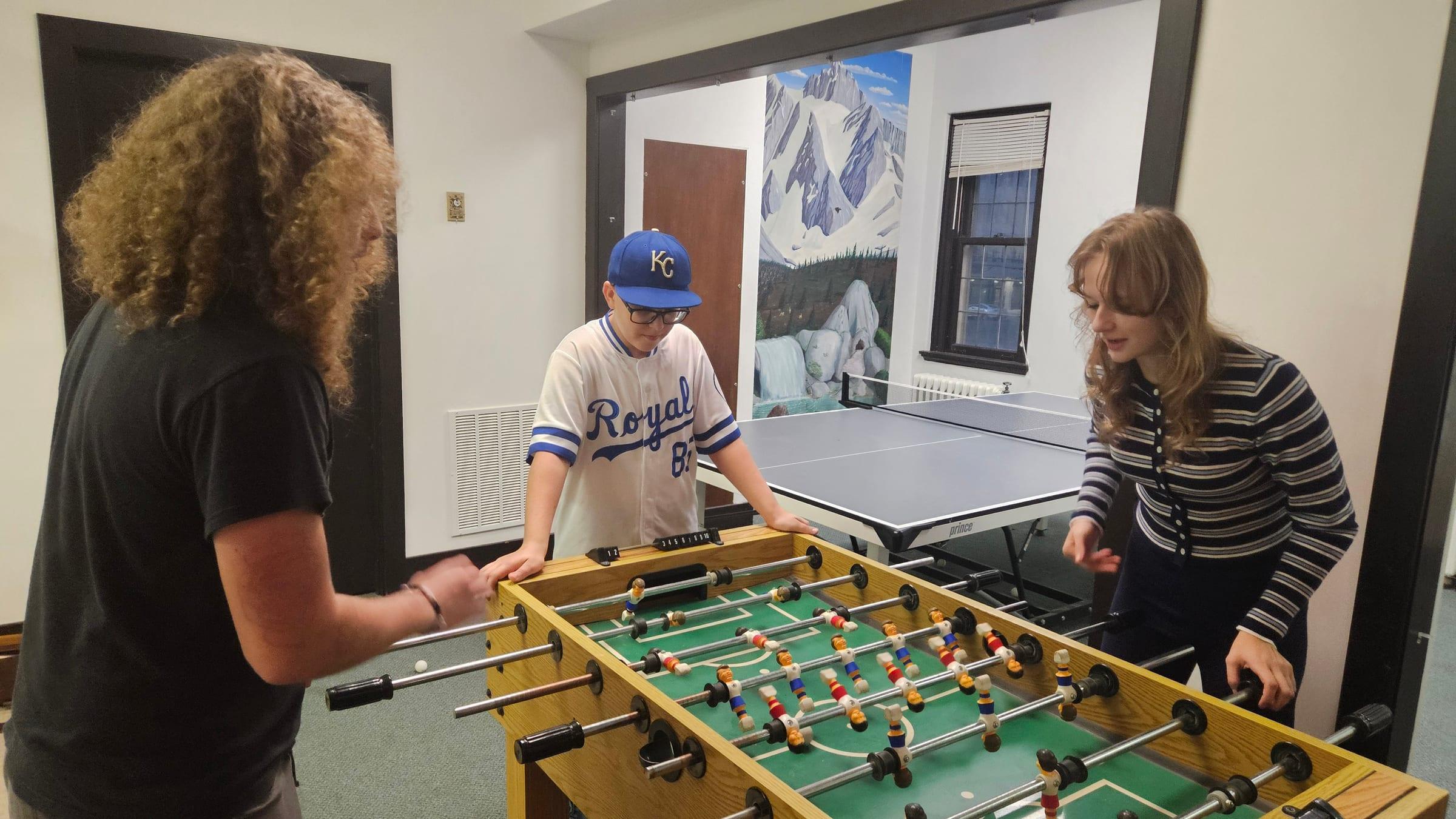 Three teenagers playing foosball in the youth room, the ping pong table behind them