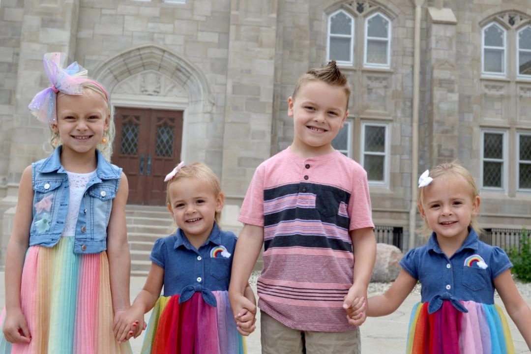 Four young children hold hands and smile, posed together outside the church's arched entrance.