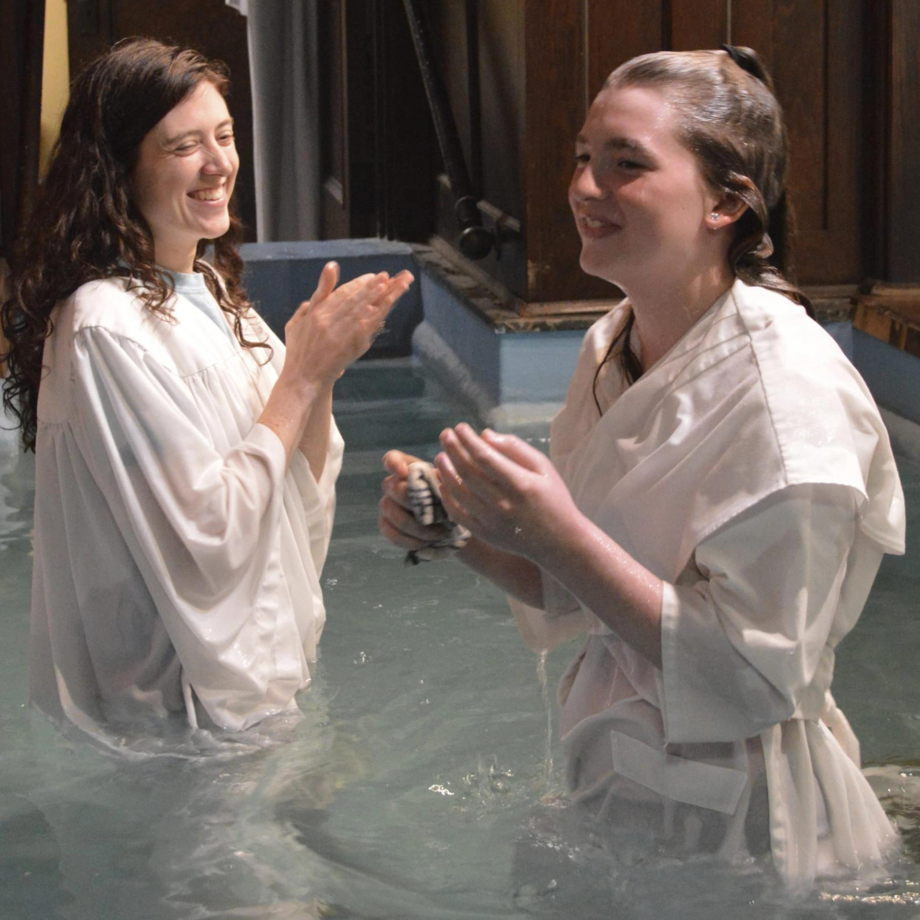 Two young women smile together in the baptismal pool, one of them just baptized