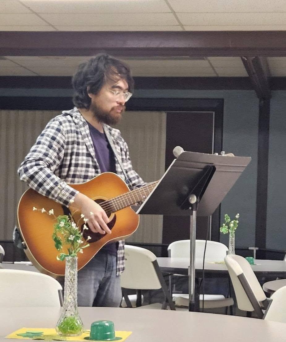 A man in a plaid shirt plays an acoustic guitar beside a music stand in the fellowship hall
