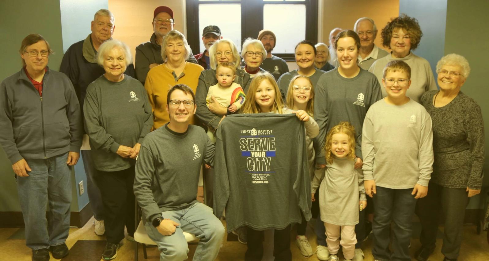 A large group of adults and children posed together indoors, some holding a team t-shirt reading Serve Your City.