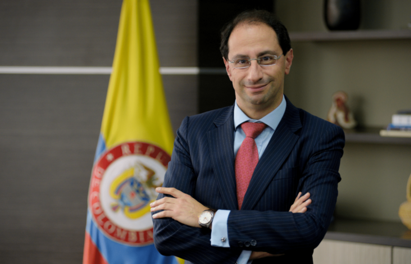 José Manuel Restrepo con traje azul y corbata roja frente a una bandera de Colombia