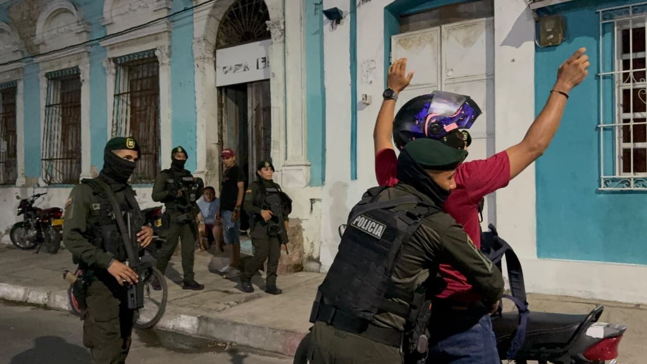 Un policía requisa a un hombre con casco y camiseta roja, mientras otros uniformados vigilan una calle.