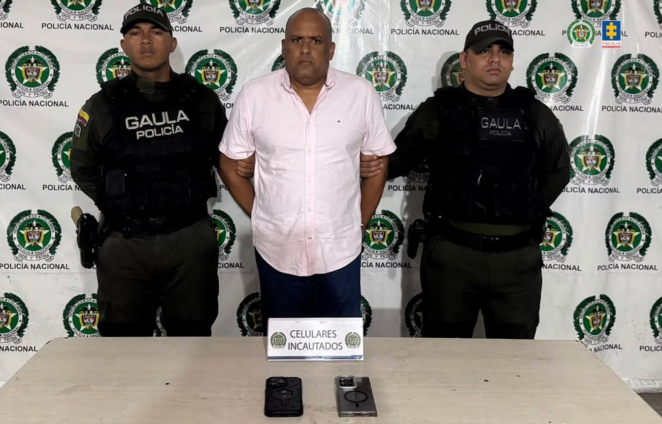 Hombre con camisa rosada custodiado por dos uniformados del Gaula; sobre una mesa hay dos celulares y un aviso de elementos incautados.
