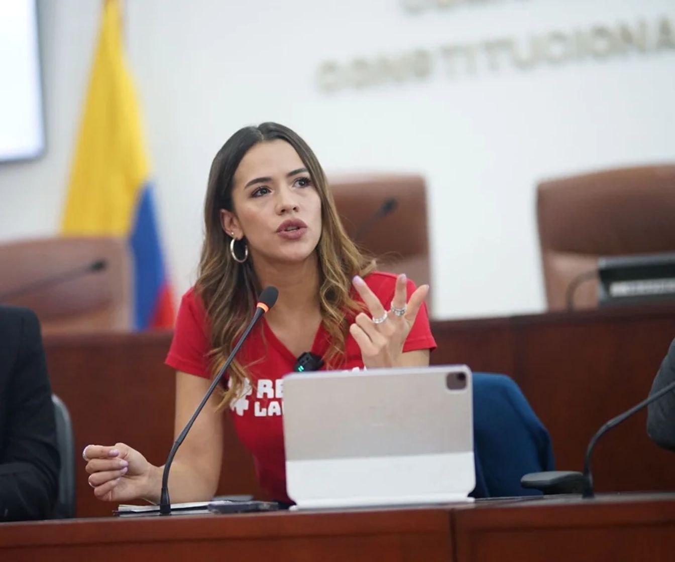 Mujer con camiseta roja habla ante un micrófono en un recinto legislativo.