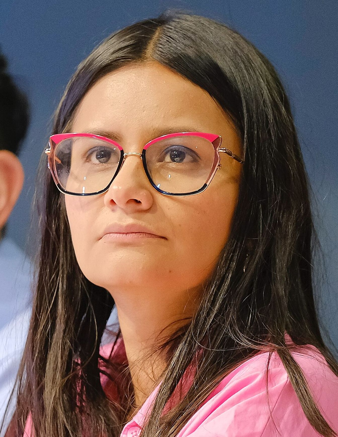 Mujer de cabello oscuro, gafas de marco rosado y camisa rosada.
