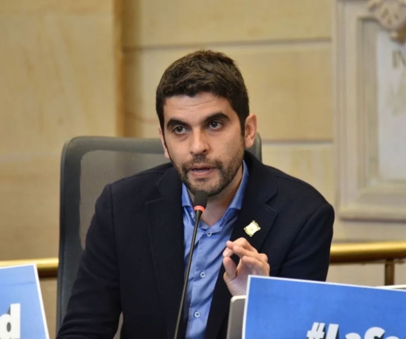 Hombre con chaqueta oscura y camisa azul interviene ante un micrófono.