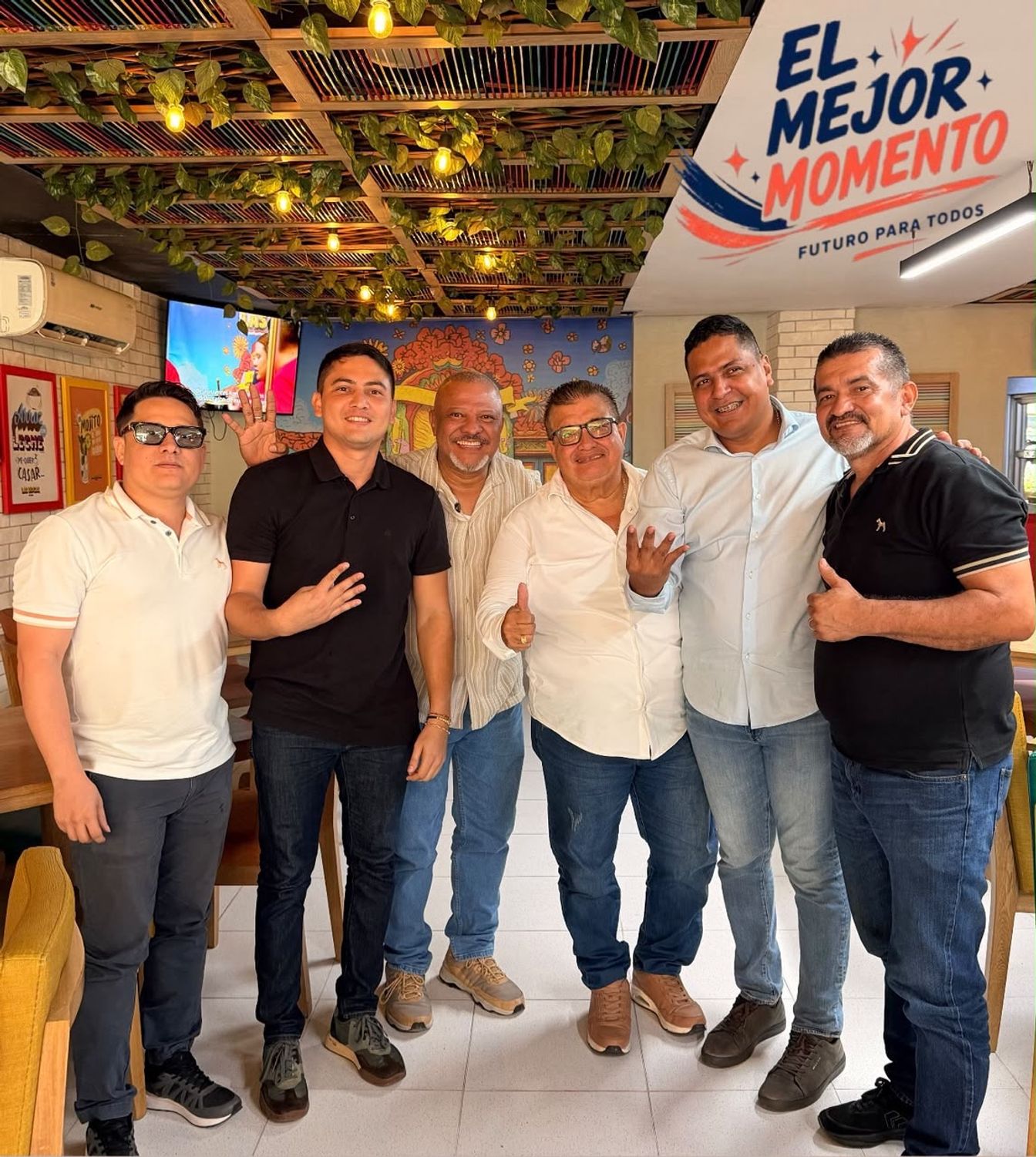 Seis hombres posan en una reunión bajo un letrero de la campaña El Mejor Momento