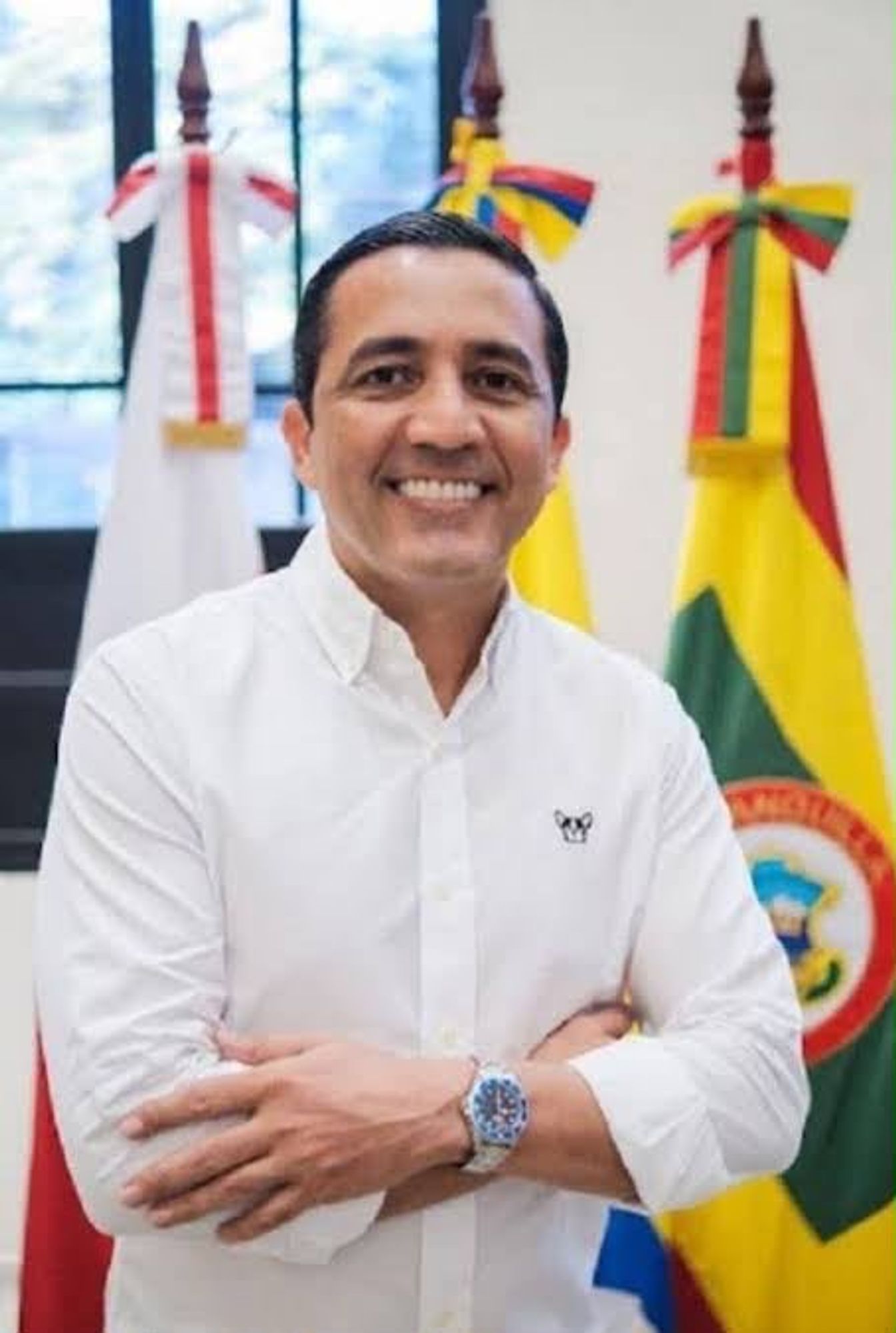 Pedro Lemus Navarro sonríe con los brazos cruzados, vestido con camisa blanca y con banderas detrás.