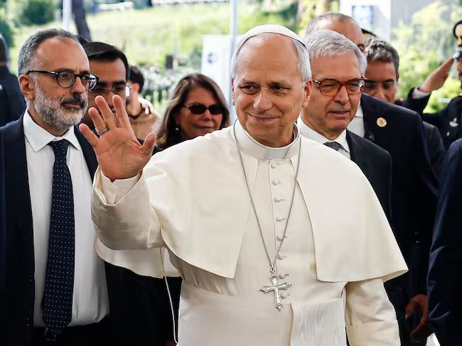 El papa León XIV saluda con la mano derecha, acompañado de varias personas.