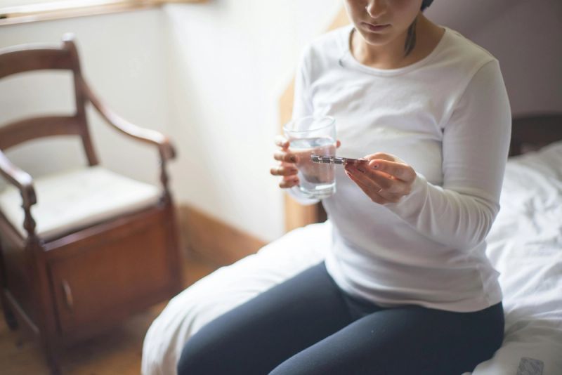 Mujer sentada en el borde de la cama, sosteniendo un vaso de agua y un blíster de medicamento