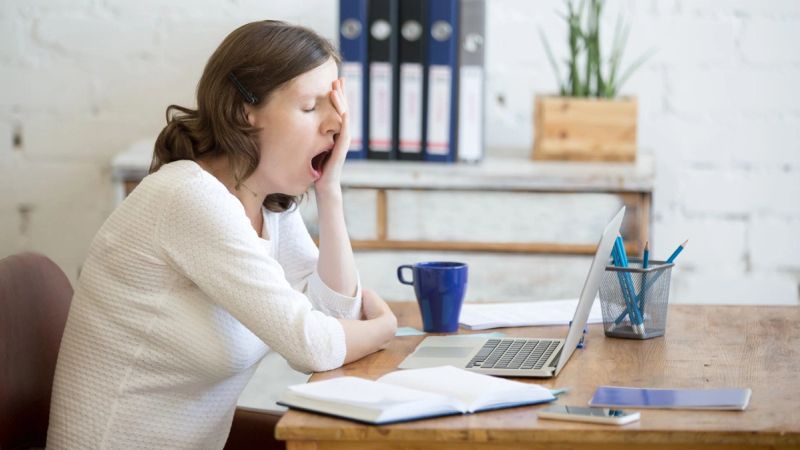 Mujer bosteza con la mano en la cara frente a su laptop en la oficina, con una taza de café al lado